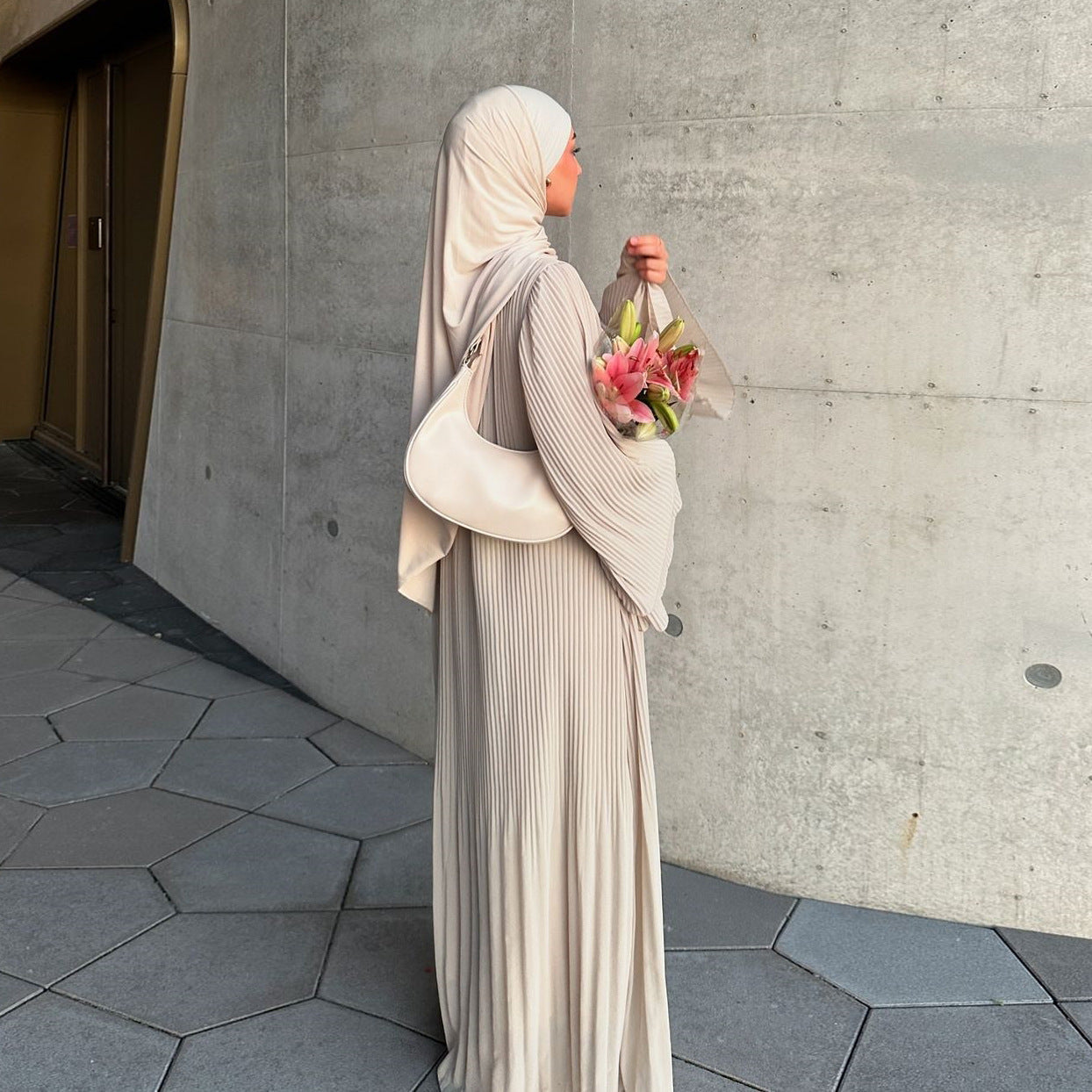 Woman in a pleated dress and hijab holding flowers, showcasing Modest Dresses collection, elegant and sophisticated style.